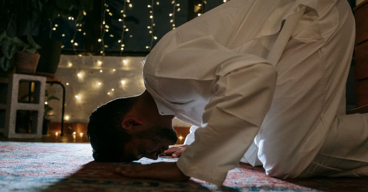 A man in traditional attire bows in prayer on a rug lit by string lights, symbolizing faith and devotion.