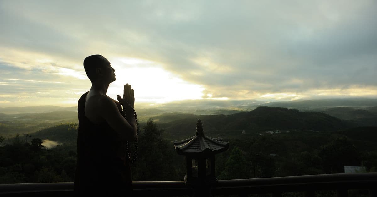 A monk in silhouette prays peacefully at sunrise with mountains in the background.